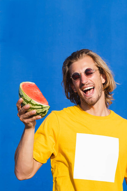 Aantrekkelijke jongeman met lang blond haar met een halve watermeloen in zijn hand glimlachend en zich vermakend op een effen blauwe achtergrond. Zomer, zon, warmte, fruit, vakantie concept. - Foto, afbeelding
