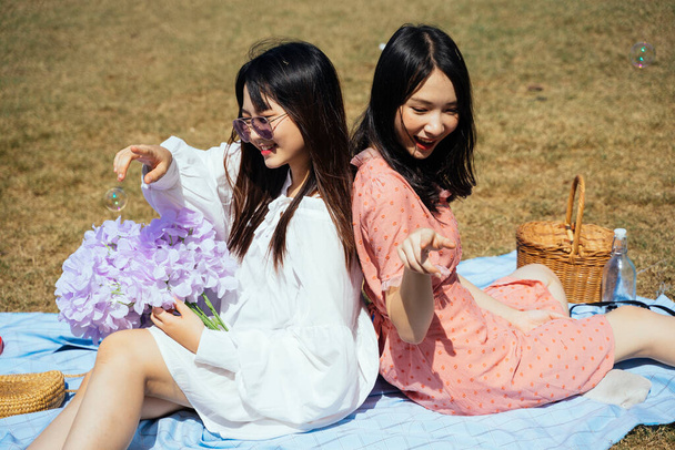 Dos chicas sentadas espalda con espalda en la alfombra en el campo, jugar la burbuja yflores púrpuras
. - Foto, Imagen