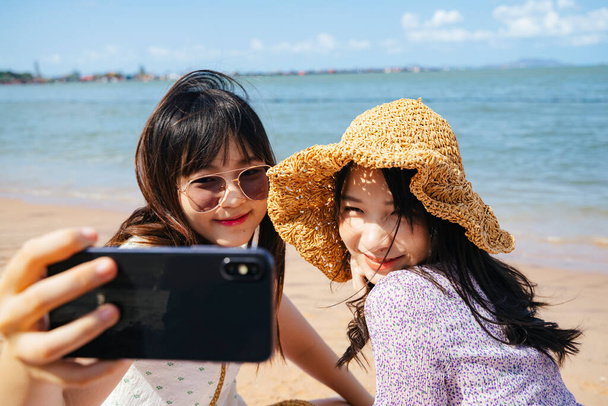 Two girls sitting on the beach at the sea in sunny day, take a selfie together and making duck face. - Photo, Image