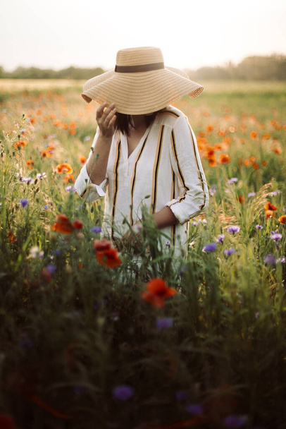 Jovem mulher em vestido de linho rústico andando entre papoula e flores de milho no campo de prado de verão na luz do pôr do sol. Vida rural lenta. Desfrutando de uma vida simples. Menina de chapéu em flores silvestres
 - Foto, Imagem