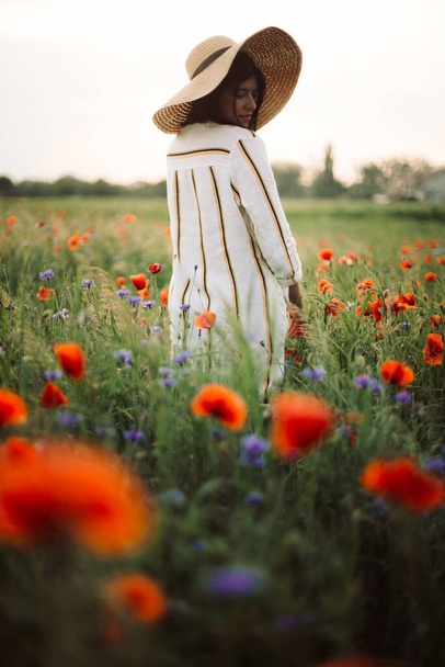 Menina elegante em chapéu andando em flores silvestres em luz do pôr do sol no prado de verão. Jovem em vestido de linho andando entre papoula e flores de milho no campo. Vida rural lenta. Desfrutando de uma vida simples
 - Foto, Imagem