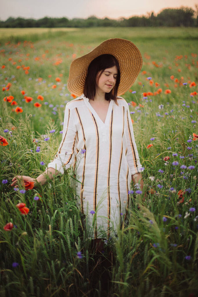 Молода жінка в сільській лляній сукні ходить серед маку і кукурудзяних квітів в літній сільській місцевості на заході сонця. Сільське повільне життя. Насолоджуйся простим життям. Дівчина в капелюсі в польових квітах
 - Фото, зображення