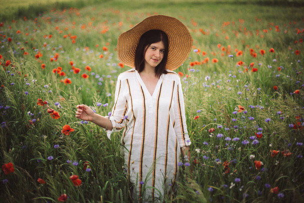 Νεαρή γυναίκα με ρουστίκ λινό φόρεμα που περπατά ανάμεσα σε παπαρούνες και άνθη καλαμποκιού στο καλοκαιρινό λιβάδι εξοχή στο φως του ηλιοβασιλέματος. Αγροτική αργή ζωή. Απολαμβάνω την απλή ζωή. Κορίτσι με καπέλο σε αγριολούλουδα - Φωτογραφία, εικόνα