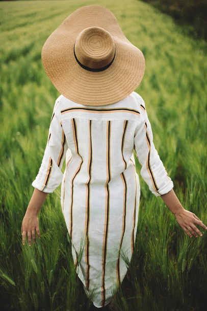 Jeune femme en robe de lin et chapeau marchant dans le champ vert de l'orge à la campagne. Fille élégante en robe rustique profiter moment paisible dans l'herbe en été. Moment rural tranquille. - Photo, image