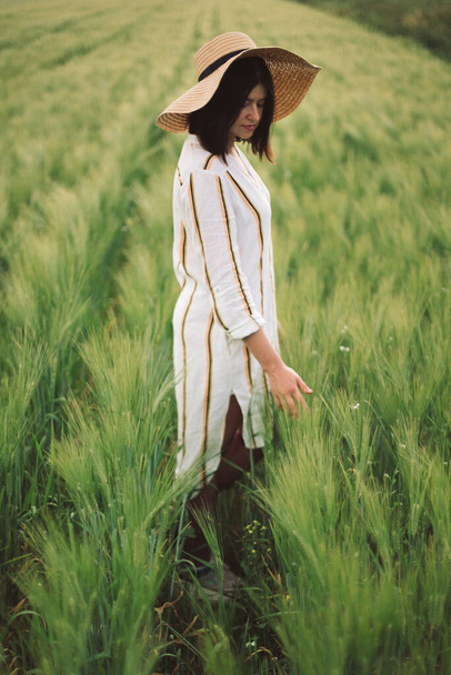 Woman in rustic dress and hat walking in green field of barley. Atmospheric authentic moment. Stylish girl enjoying peaceful evening in countryside. Rural slow life - 写真・画像