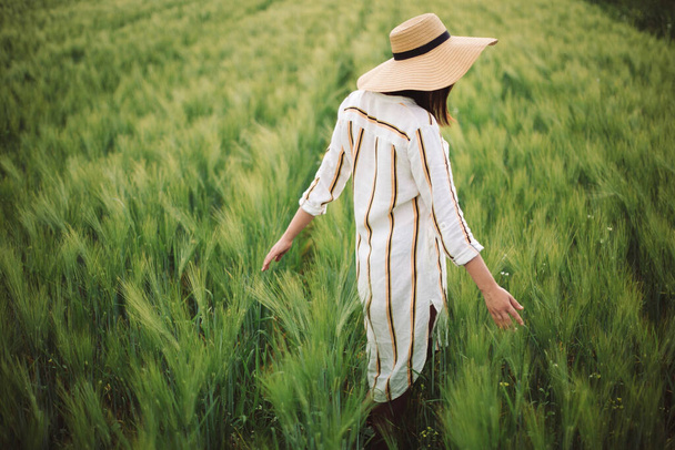 Jonge vrouw in linnen jurk en hoed wandelen in groen veld van gerst op het platteland. Stijlvol meisje in rustieke jurk genieten van rustig moment in gras in de zomer. Rust uit op het platteland. Kopieerruimte - Foto, afbeelding