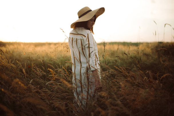 Frau in rustikalem Kleid und Hut, die in Wildblumen und Kräutern im goldenen Licht des Sonnenuntergangs auf der Sommerwiese wandelt. Stimmungsvoller authentischer Moment. Stilvolles Mädchen genießt Abend auf dem Land. - Foto, Bild
