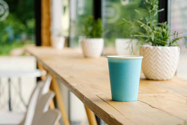 Eine blaue Pappbecher mit heißem, frischem Kaffee steht auf einem Holztisch in einem Café. Das Innere des Cafés mit einem Ripsalis-Kaktus (Zweig) und anderen grünen Pflanzen. Der Blick aus dem Café. - Foto, Bild