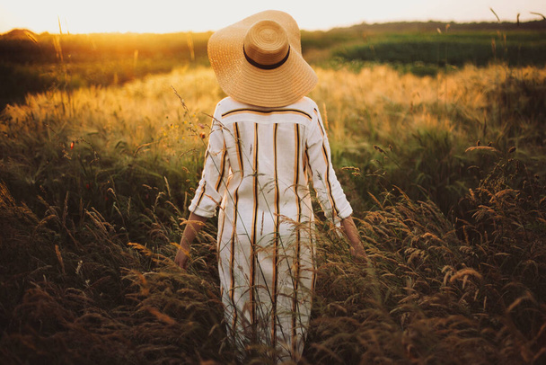 Γυναίκα σε ρουστίκ φόρεμα και καπέλο περπάτημα σε αγριολούλουδα και βότανα στο ηλιοβασίλεμα χρυσό φως στο θερινό λιβάδι. Ατμοσφαιρική αυθεντική στιγμή. Κομψό κορίτσι απολαμβάνοντας το βράδυ στην ύπαιθρο. - Φωτογραφία, εικόνα