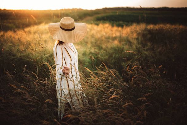 Vrouw in rustieke jurk en hoed wandelen in wilde bloemen en kruiden in zonsondergang gouden licht in de zomer weide. Stijlvol meisje geniet van de avond op het platteland. Platteland langzaam leven. Kopieerruimte - Foto, afbeelding
