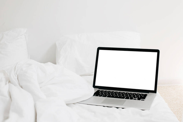 Laptop with blank white screen on white cozy bed in sunny light, copy space. Shopping online from bed. Working online from home. Mockup. Freelance concept. Staying home. Blog template - Photo, Image