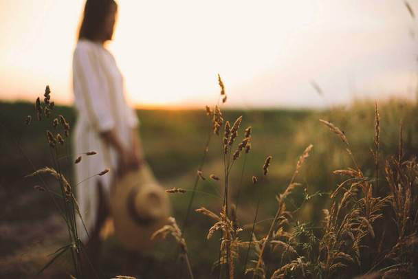 Kräuter und Gräser im Sonnenuntergang auf dem Hintergrund einer verschwommenen Frau auf der Sommerwiese. Wildblumen aus nächster Nähe in warmem Licht und rustikale Mädchen, die sich abends in der Natur entspannen. Ruhiger Moment - Foto, Bild