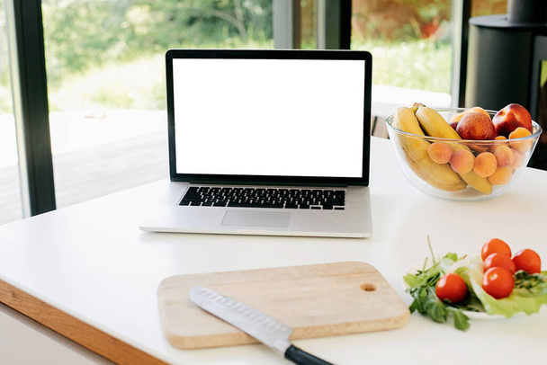 Cooking home concept. Food blog. Laptop with blank white screen and vegetables, knife and fruits on countertop in modern kitchen with big windows, copy space. Culinary courses or recipe online. - Photo, Image
