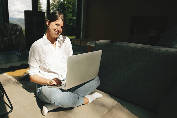 Fiatal nő használja laptop és mosolygós, online vásárlás vagy dolgozik online otthonról. Szabadúszó és szabadúszó. Boldog elegáns lány ül a padlón laptoppal napsütésben - Fotó, kép