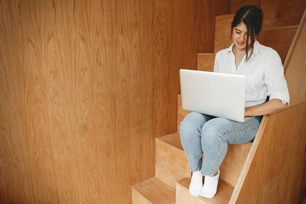 Pracuję online. Szczęśliwa stylowa dziewczyna za pomocą laptopa, siedzi na drewnianych schodach z filiżanką kawy. Młoda kobieta zakupy online lub pracy online z domu i uśmiechnięty. Wolny strzelec - Zdjęcie, obraz