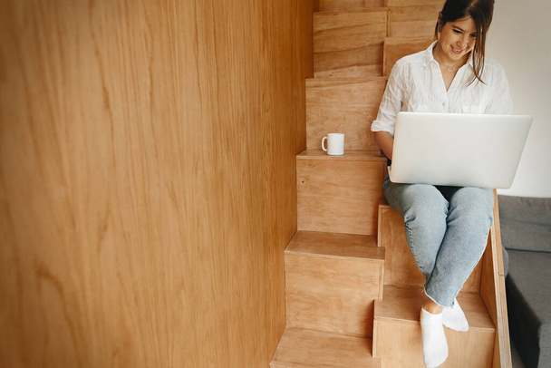 Счастливая стильная девушка с ноутбуком, сидящая на деревянной лестнице с чашкой кофе. Молодая случайная женщина делает покупки онлайн или работает онлайн из дома и улыбается. Сиди дома. Фрилансер
 - Фото, изображение