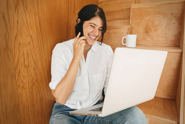 Молода випадкова жінка купує онлайн або працює в Інтернеті з дому і посміхається. Щаслива стильна дівчина спілкується по телефону і використовує ноутбук, сидячи на дерев'яних сходах в сучасній кімнаті. Залишайся вдома. Фріланс
 - Фото, зображення