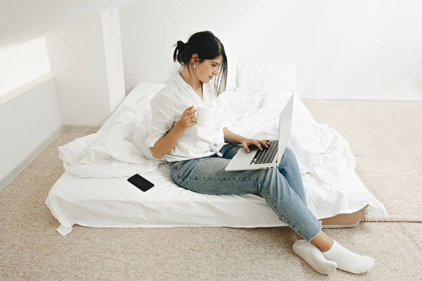 Heureuse fille élégante assise sur un lit blanc avec ordinateur portable et tenant une tasse de café. Jeune femme avec ordinateur portable, navigation, achats en ligne ou de travail en ligne. Reste à la maison. Freelance - Photo, image