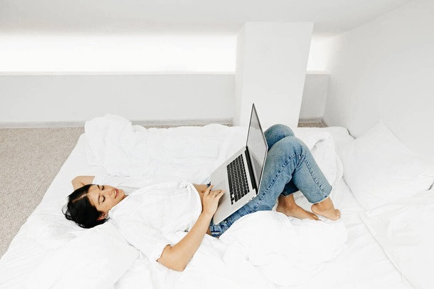 Happy stylish girl using laptop, lying on bed in white modern room. Young casual woman shopping online or working online from home and smiling. Stay home. Freelance - Фото, изображение