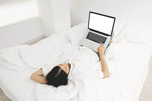 Boldog elegáns lány fekszik az ágyban laptoppal üres képernyő fehér modern szobában. Fiatal nő keres üres kijelző, online vásárlás vagy dolgozik online otthonról sablon. Szabadúszó - Fotó, kép