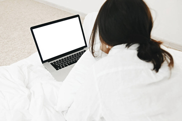 Fille élégante couchée sur le lit avec ordinateur portable avec écran blanc dans la chambre moderne blanche. Jeune femme regardant l'affichage vide, faisant des achats en ligne ou travaillant en ligne à partir du modèle à la maison. Freelance - Photo, image