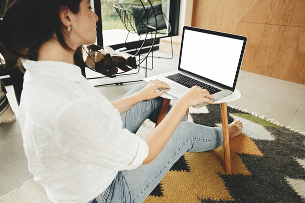 Elegante chica hipster utilizando el ordenador portátil con pantalla en blanco, sentado en el suelo en la habitación moderna. Joven mujer de compras o de trabajo en línea desde casa se burlan. freelance y freelancer
 - Foto, imagen