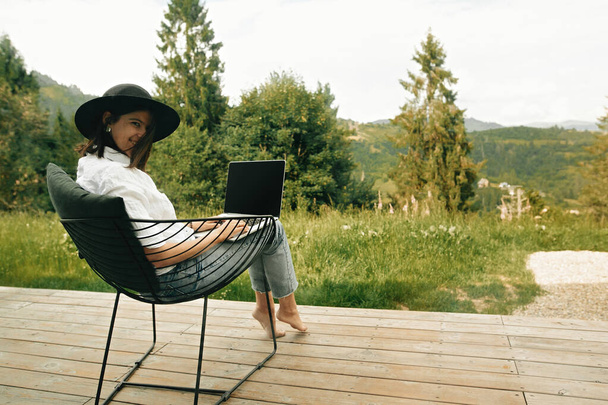 Hipster tyttö kannettavan istuu puinen kuisti kaunis näkymä metsään ja vuorille. Tyylikäs nuori nainen hattu kannettavan tietokoneen avulla, ostoksia tai työskentelevät verkossa ulkopuolella. Freelance ja freelancer - Valokuva, kuva