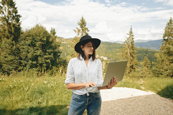 Hipster girl using laptop on background of beautiful view on woods and mountains. Stylish young woman in hat blogging, shopping or working online outside. Travel and Freelance. - Фото, изображение