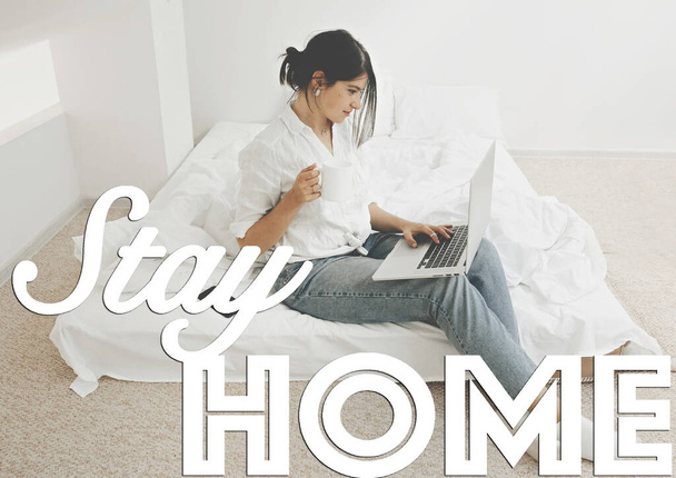 Stay home text. Happy stylish girl sitting on white bed with laptop and holding cup of coffee. Stay home save lives. Isolation in home to prevent epidemic - Photo, Image