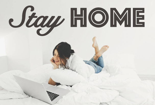 Stay home text. Happy stylish girl looking at laptop, lying on bed in white modern room, and smiling. Freelance. Stay home save lives. Isolation in home to prevent epidemic - Photo, Image