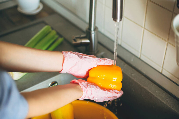 Manos en guantes sosteniendo pimienta, lavando verduras durante la epidemia de virus. Mujer en manos rosadas lavando verduras frescas, preparándose para cocinar comida en la cocina moderna
 - Foto, Imagen