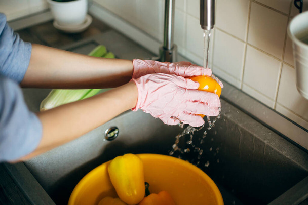 Lavando verduras. Las manos en los guantes lavando pimienta bajo el chorro de agua en el fregadero durante la epidemia de virus. Mujer en manos rosadas limpiando verduras frescas, preparándose para cocinar comida en la cocina moderna
 - Foto, Imagen