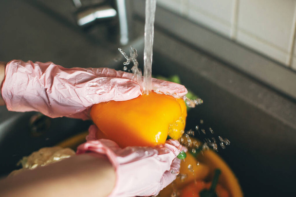 Mycie warzyw. Ręce w rękawiczkach myjących pieprz pod strumieniem wody w zlewie podczas epidemii wirusa. Kobieta w różowych rękach sprzątanie świeżych warzyw, przygotowanie do gotowania posiłku w nowoczesnej kuchni - Zdjęcie, obraz