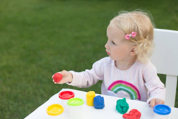Blond toddler girl playing modeling clay in the garden in sunny summer day - Foto, Bild