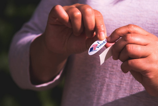 American woman peeling sticker off of backing to place on clothes to communicate to others that she had voted after leaving polling location - Photo, Image
