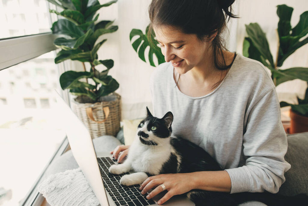 彼女の猫とラップトップで働いているカジュアルな女の子は、枕や植物と現代的な部屋に一緒に座っています。検疫中にかわいい猫の助けの所有者。快適な仲間。家にいて - 写真・画像