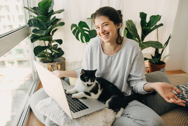 Cute cat helping owner during quarantine, loyal companion. Casual girl working on laptop with her cat, sitting together in modern room with pillows and plants. Home office.  Stay home stay safe - Photo, image