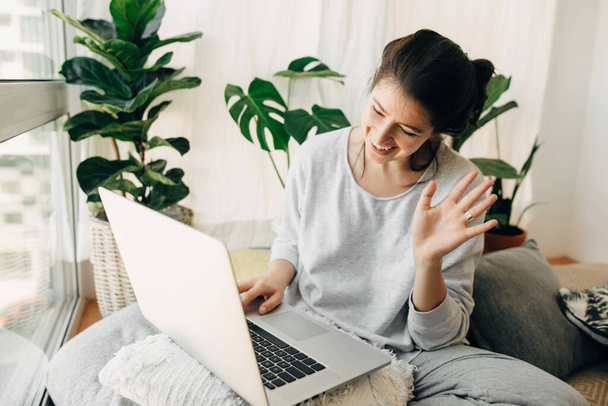 幸せな女の子ノートパソコンにハイ手を振って、ビデオ、家族や友人とチャット、枕や植物と近代的な部屋に座っている。隔離中の通信にラップトップを使用している若いカジュアルな女性。内務省 - 写真・画像