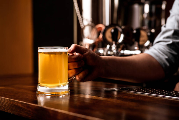 Hrnek piva pro klienta. Barman klade lehký ležák na dřevěný hnědý pult baru - Fotografie, Obrázek