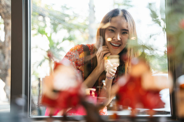 Joven mujer asiática en vestido rojo mirando cosas detrás de la ventana de compras en la calle con expresión alegre
. - Foto, Imagen