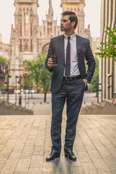 Pauza na kafe. Sebevědomý mladý muž v plném obleku drží šálek kávy a dívá se stranou, zatímco stojí venku s městskou krajinou v pozadí. - Fotografie, Obrázek