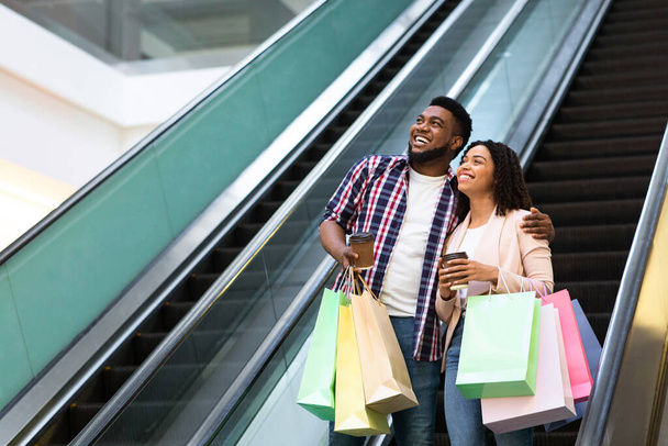 Feliz pareja negra bajando por la escalera mecánica después de compras exitosas en el centro comercial
 - Foto, Imagen