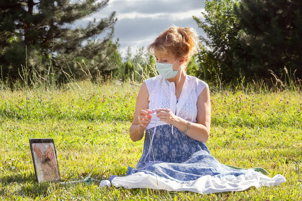 kobieta z maską medyczną na twarzy siedzi na trawie w parku dziergając wełniane ubrania na drutach dziewiarskich i obserwuje lekcje lub uczy na drutach przez laptopa. Kształcenie na odległość - Zdjęcie, obraz