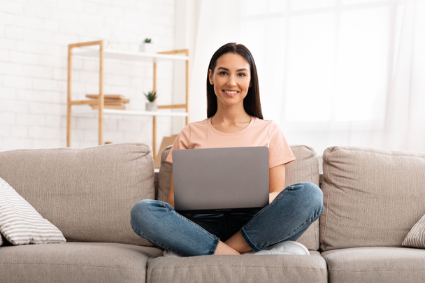 Kobieta korzystająca z laptopa siedzi na kanapie, patrząc w kamerę - Zdjęcie, obraz