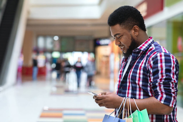 Aplikacja ze zniżkami. Uśmiechnięty czarny człowiek za pomocą smartfona w centrum handlowym - Zdjęcie, obraz