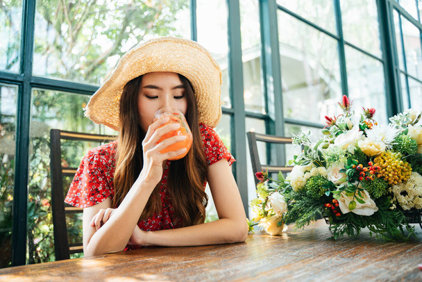 Young asian thai woman on red dress wearing straw hat drinking ice cold juice at green cafe. - Foto, Imagem