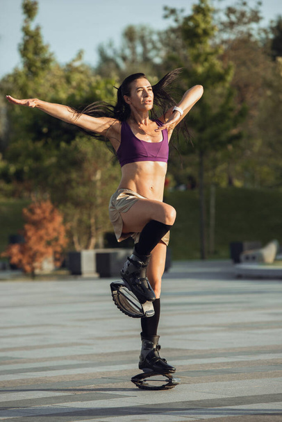 Beautiful woman in sportswear jumping in a kangoo jumps shoes at the street on summers sunny day - Fotoğraf, Görsel