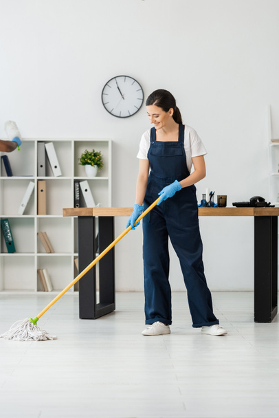 Smiling cleaner washing floor with mop near african american colleague in office - Foto, Imagem