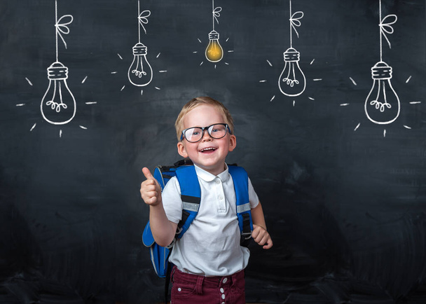 Back to school. Funny little boy in glasses pointing up on blackboard. Child from elementary school with book and bag. Education. Bulb with light on blackboard. - Photo, Image