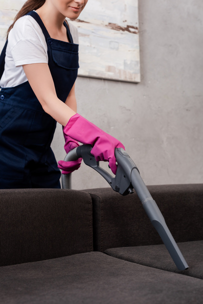 Cropped view of cleaner in rubber gloves cleaning upholstery of couch with vacuum cleaner - Foto, Imagen